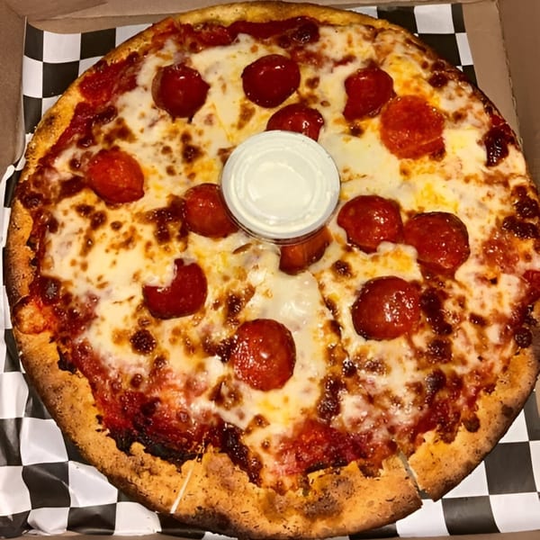 A pepperoni pizza with melted cheese and a small cup of dipping sauce in a box.