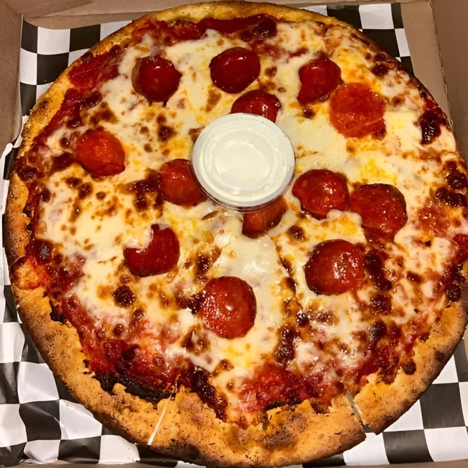 A pepperoni pizza with melted cheese and a small cup of dipping sauce in a box.