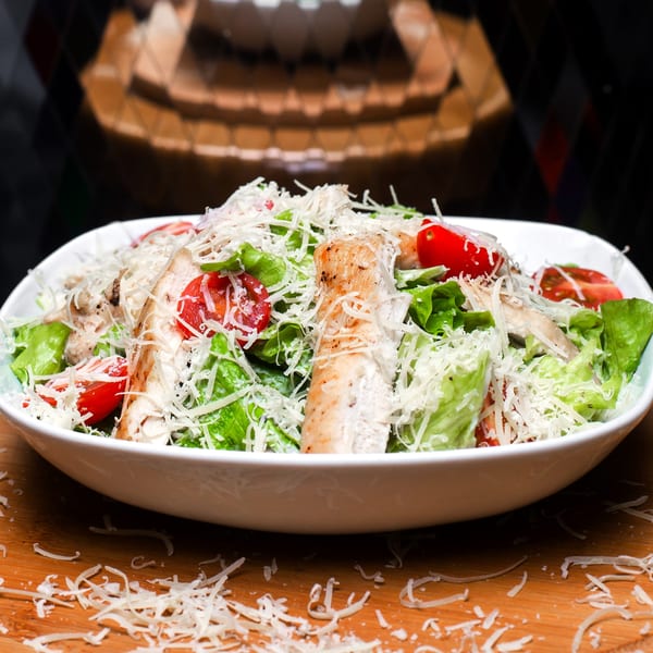 A Caesar salad with grilled chicken, cherry tomatoes, lettuce, and shredded cheese in a white bowl.